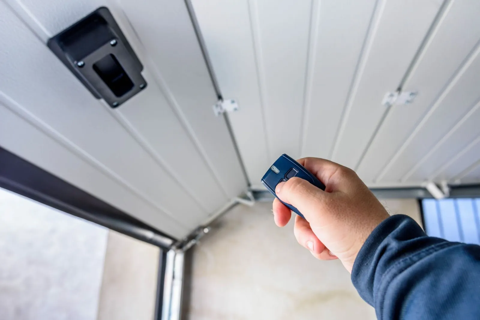 A person in a blue sleeve holds a remote to open a garage door, viewed from inside the garage with a wall-mounted opener visible.