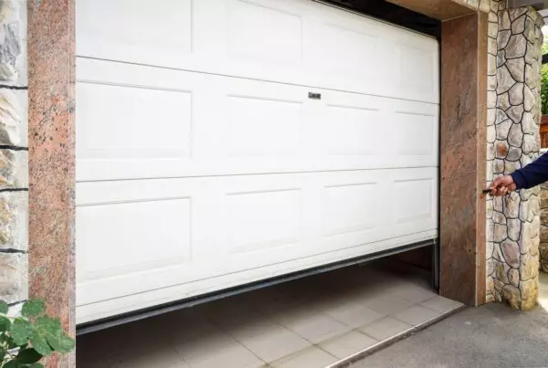 A person in a blue sleeve opens a white garage door with a remote, framed by stone and granite walls, with a plant visible on the left.