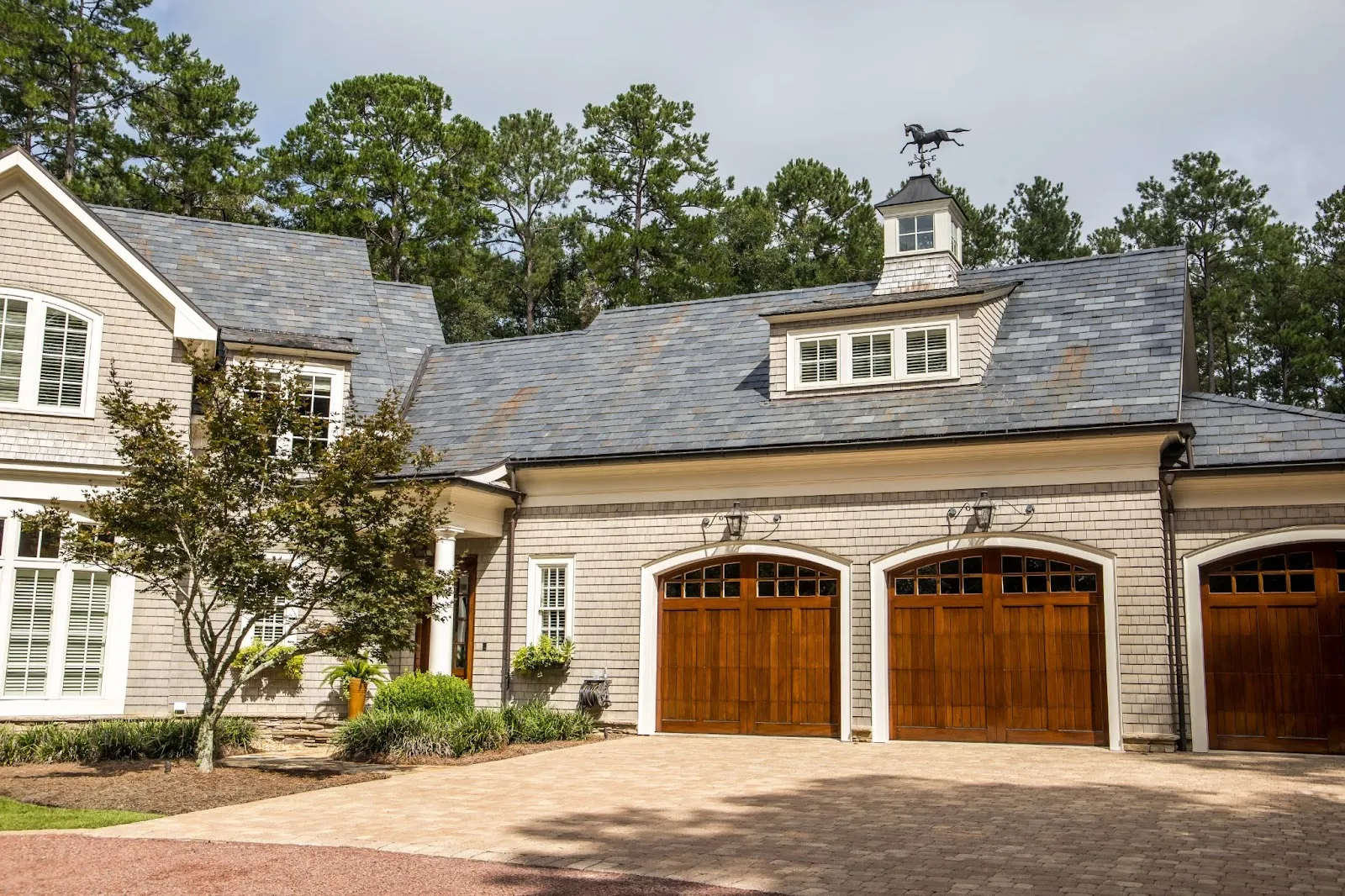 A two-story house with a slate roof, wooden double garage doors, and shingle siding, surrounded by trees and a paver driveway.